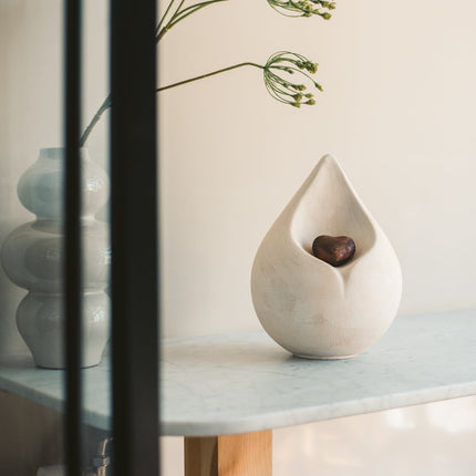 Biologische urn in de vorm van een druppel met een los hartje erin. De urn  heeft een witte kleur en hartje heeft een bronze en zwarte kleur. De urn staat gestationeerd op een rond houten tafeltje met een marmeren blad met naast zijn rechterzijde een vaas met een plant erin.