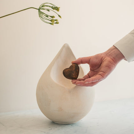 Biologische urn in de vorm van een druppel met een los hartje erin. De urn  heeft een witte kleur en hartje heeft een bronze en zwarte kleur.  Het hartje word getoond door een hand hand vlak voor de urn. De urn staat gestationeerd op een blad met een plant erboven.