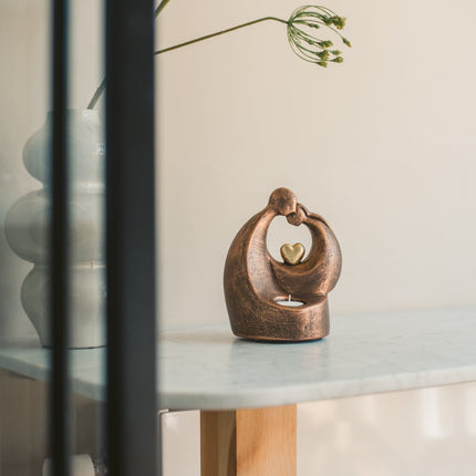 De urn is gemaakt van keramiek en heeft de kleur brons met zwart. De urn heeft een goud hartje in het midden met aan de voorkant een plek voor een kaarsje. De urn staat gestationeerd op een tafel met een blauw marmeren blad. Naast de urn staat een plant.