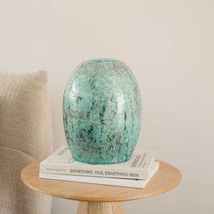 De urn is gemaakt van keramiek en heeft de kleur groen. De urn is open te maken vanaf de bovenkant. De urn staat gestationeerd op twee boeken op een houten tafeltje naast de bank.