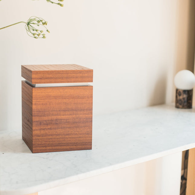 De urn is gemaakt van houtlaminaat en heeft de vorm van een kubus. De kleur die de urn heeft is bruin met een witte rand. De urn staat gestationeerd op een tafel met een wit marmeren blad boven de urn hangen bloemen. 